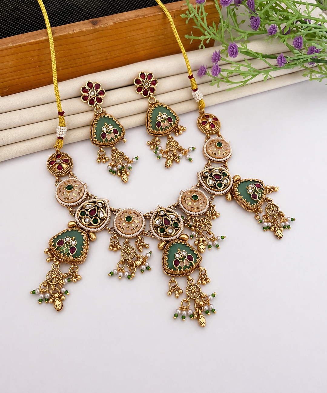 Ornate gold necklace with green and red accents on a white surface with plants.
