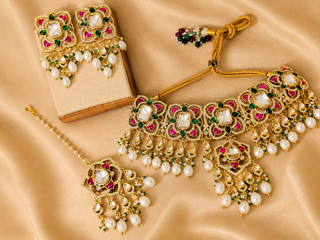 Layered polki-inspired detailing with ruby and emerald-toned accents, pearl drops, and matching earrings on beige background.