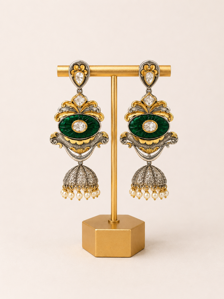 Ornate earrings with green gemstones and gold accents on a stand against a beige background