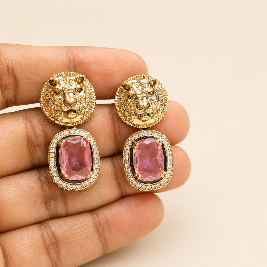 Gold lion head earrings with pink gemstones held in a hand against a beige background
