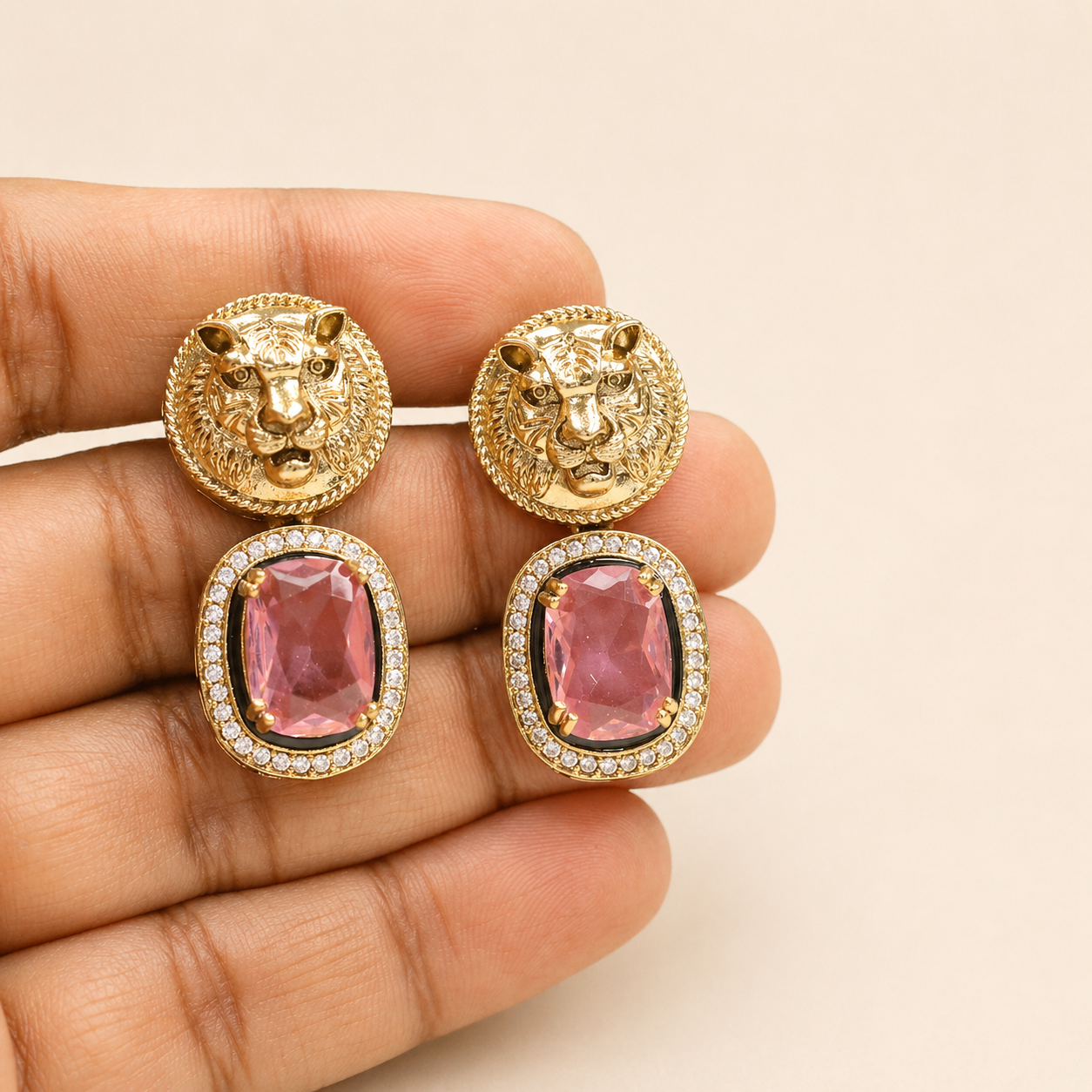 Gold lion head earrings with pink gemstones held in a hand against a beige background